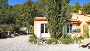 Una casa rústica con entrada de grava, un pequeño espacio exterior con mobiliario y un árbol grande al frente.