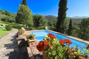 A swimming pool with a view of a mountainous landscape, surrounded by greenery and a few lounge chairs.