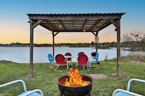 A wooden gazebo with a fire pit in the foreground, surrounded by outdoor seating and a view of a lake.