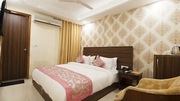 A hotel room with a large bed, a wooden headboard, a nightstand, a desk, a chair, a wardrobe, an air conditioning unit, and a ceiling fan.