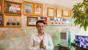 A hotel reception area with framed photographs on the wall, a computer monitor displaying images, and a plant in the foreground. - Cambodia Eren Hotel (Phnom Penh)