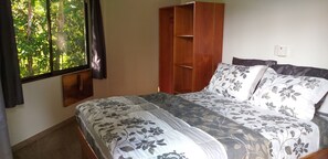 A bedroom with a bed, a wooden wardrobe, a window with curtains, and a wall-mounted light switch.
