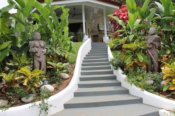 A staircase leading to a covered porch with statues and lush tropical plants.