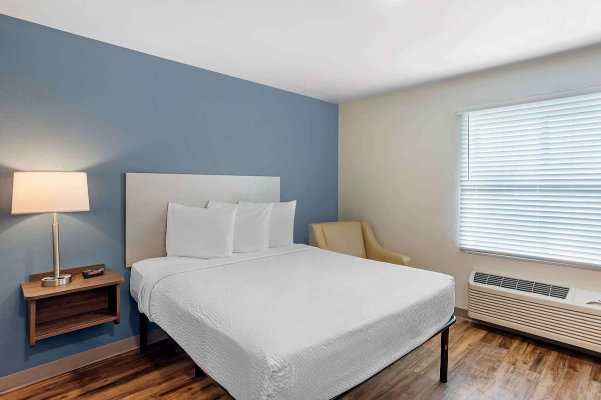 A neatly arranged hotel room with a bed, bedside lamp, chair, and air conditioning unit.