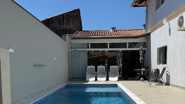 A rectangular swimming pool with a tiled edge, surrounded by a white wall and a building with a tiled roof. There are two potted plants in the foreground.