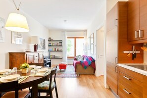 A modern kitchen with wooden cabinets, a dining area with a table set for four, and a cozy living room with a sofa and a colorful blanket.