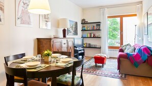 Un coin repas avec une table dressée pour quatre personnes, un téléviseur posé sur un meuble, une bibliothèque et une fenêtre avec des rideaux.