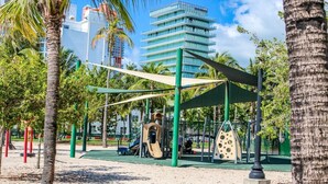 Un parque infantil con toboganes y estructuras para escalar, rodeado de palmeras y un edificio al fondo.