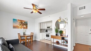 Una sala de estar moderna con un sofá de cuero oscuro, una mesa de comedor de madera con sillas, un ventilador de techo, una cocina con armarios blancos, un microondas y un espejo redondo sobre una consola.