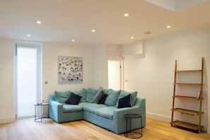 A modern living room with a teal sofa, a small side table, a wooden ladder shelf, and a bird painting on the wall.