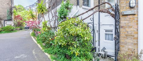 Une rue résidentielle avec une maison blanche, un portail noir et un jardin où poussent différentes espèces de plantes.