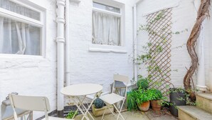 A white-walled room with a small round table and two chairs, a potted plant, and a climbing vine.
