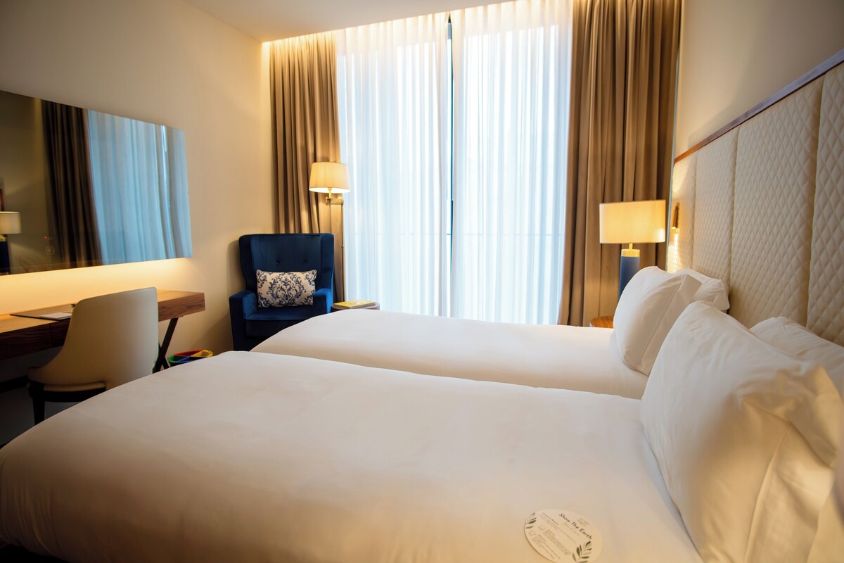 A hotel room with two beds, a desk, a chair, and a large window with curtains.