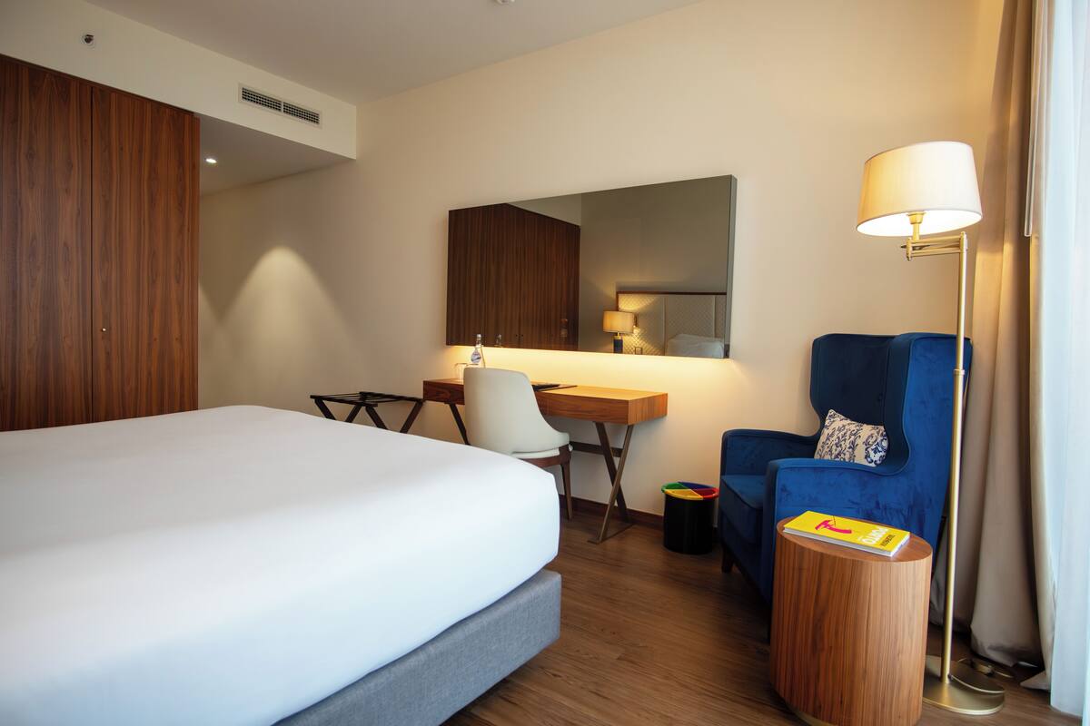 A hotel room with a large bed, a desk with a chair, a blue armchair, a small round table with a book, and a floor lamp.