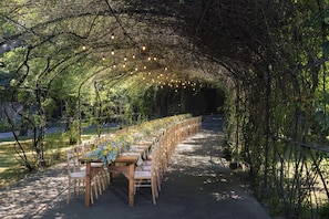 Un área de comedor al aire libre con mesas y sillas dispuestas bajo un dosel de árboles, con luces colgantes.