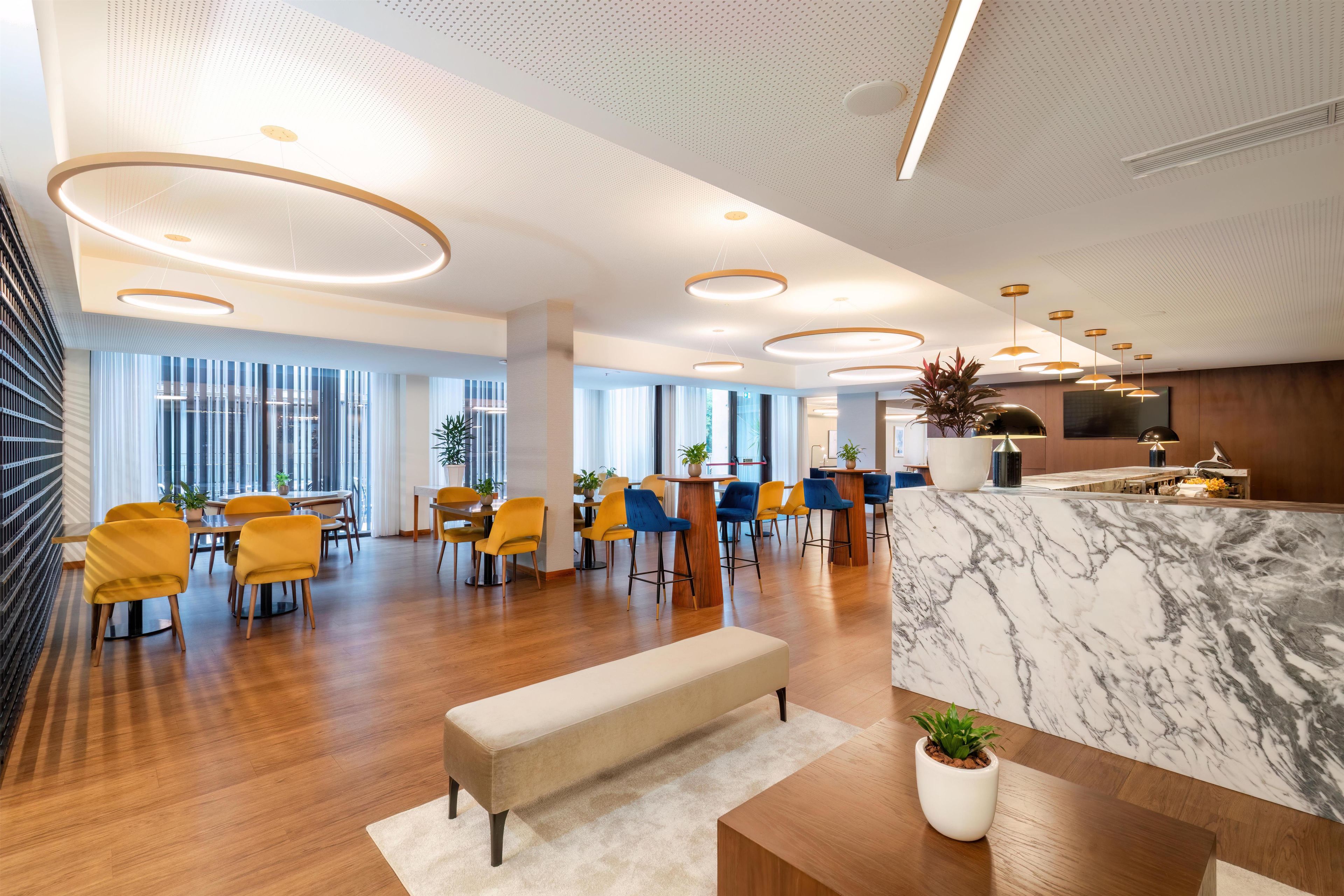 A modern hotel lobby with a marble reception desk, wooden flooring, and yellow chairs.