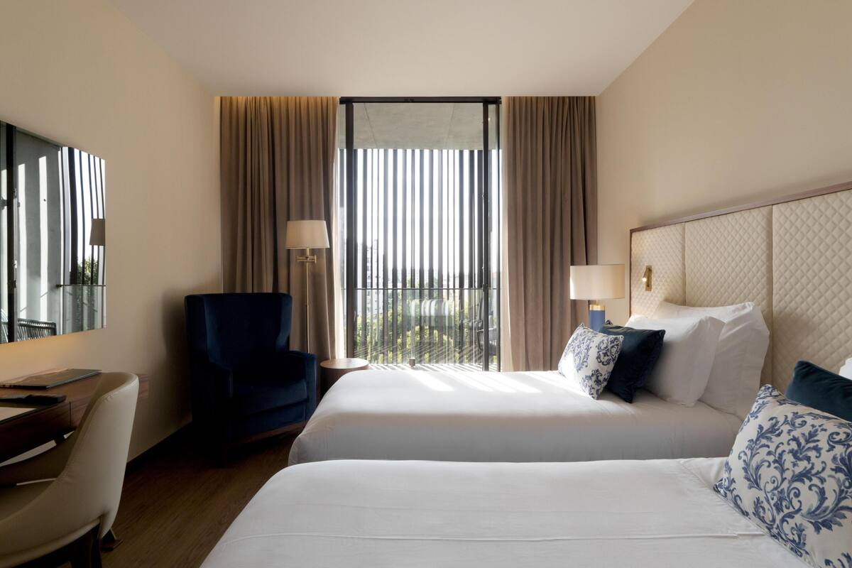 A hotel room with two beds, a desk, a chair, and a large window with curtains.