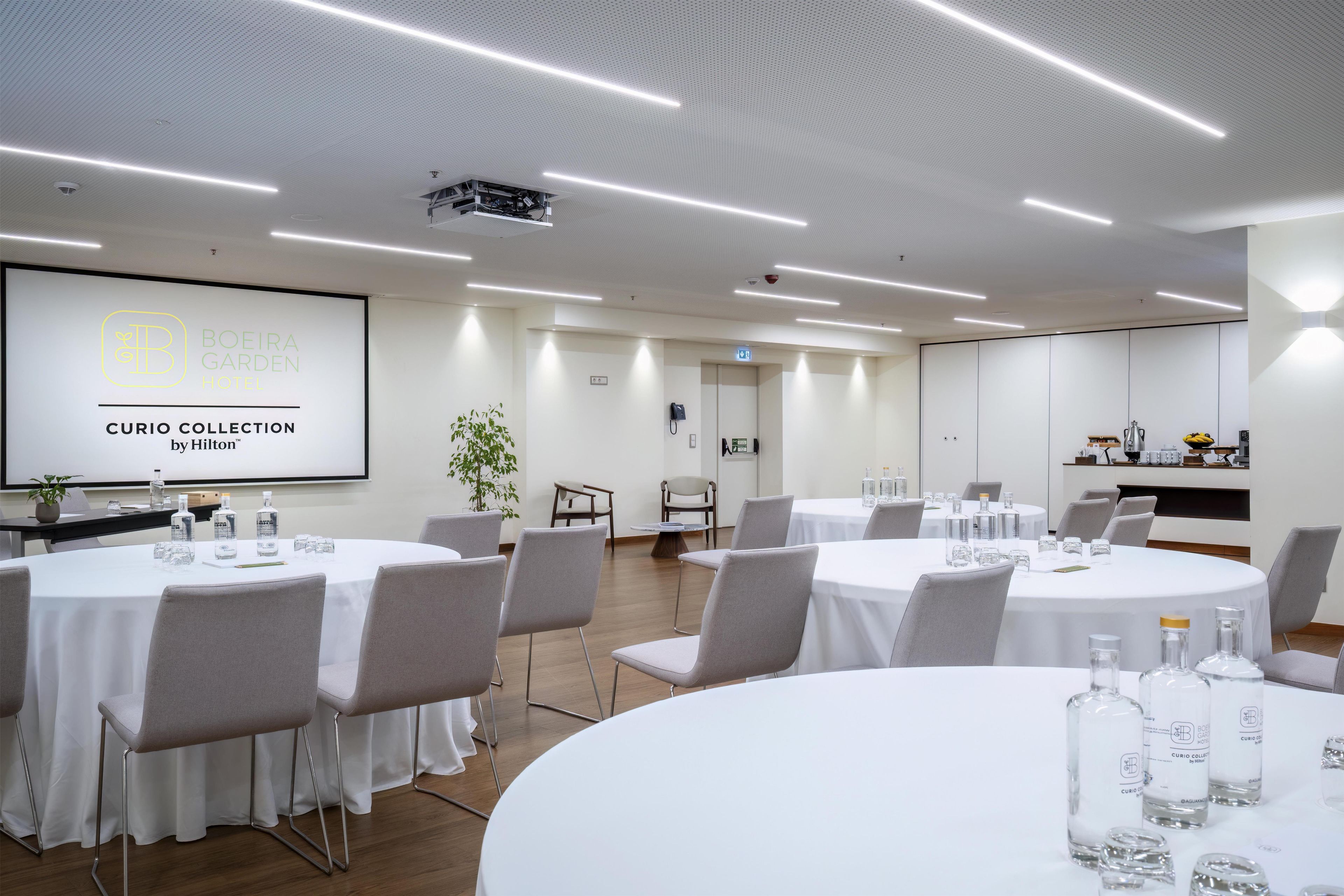 Un salón de conferencias con mesas blancas, sillas grises y una pantalla de proyector mostrando 'BOEIRA GARDEN HOTEL CURIO COLLECTION by Hilton'.