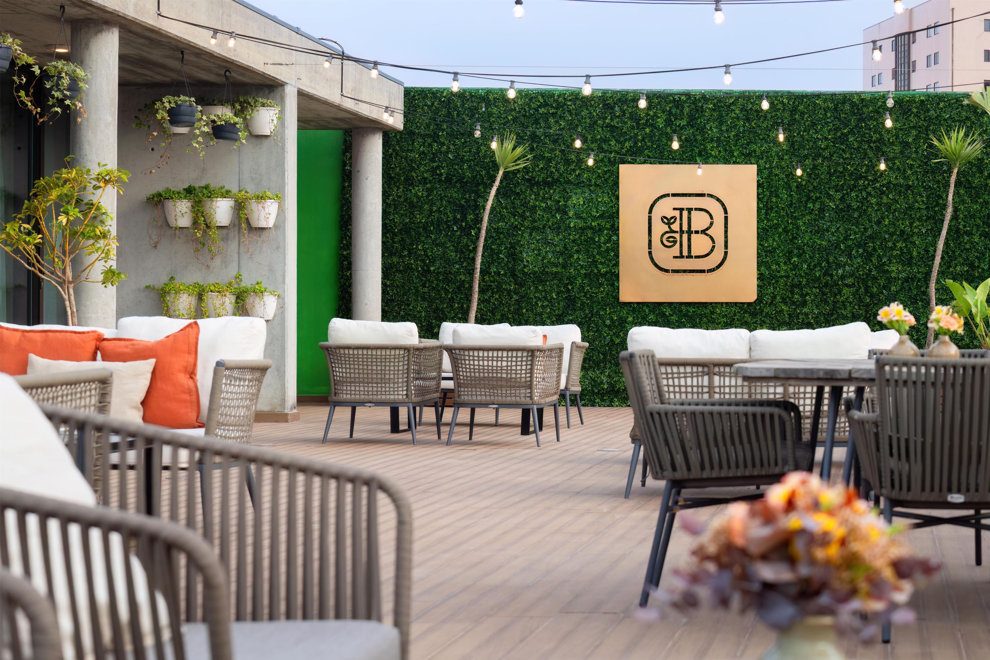 An outdoor seating area with wicker furniture, potted plants, and a green wall with a logo.