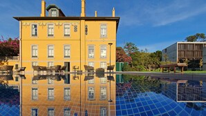 Un edificio amarillo con reflejo en una piscina, mesas al aire libre y una estructura moderna cerca.