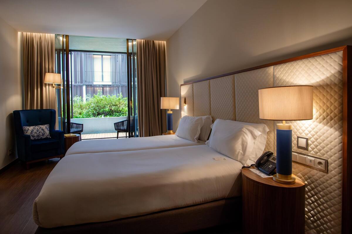 A hotel room with two beds, a desk, a chair, and a view of greenery outside.