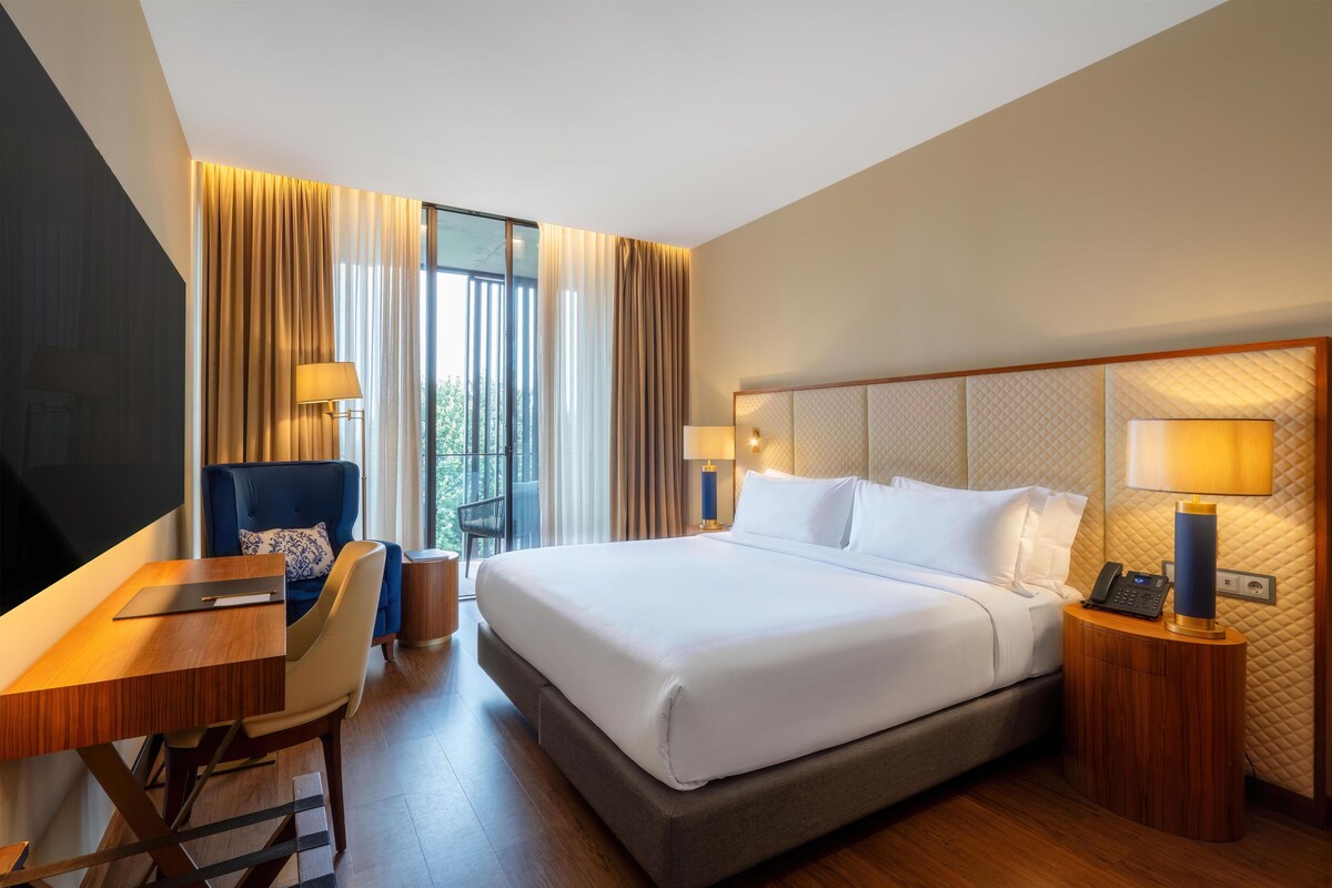 Une chambre d’hôtel avec un lit, un bureau, une chaise, une table de chevet et une fenêtre avec des rideaux.