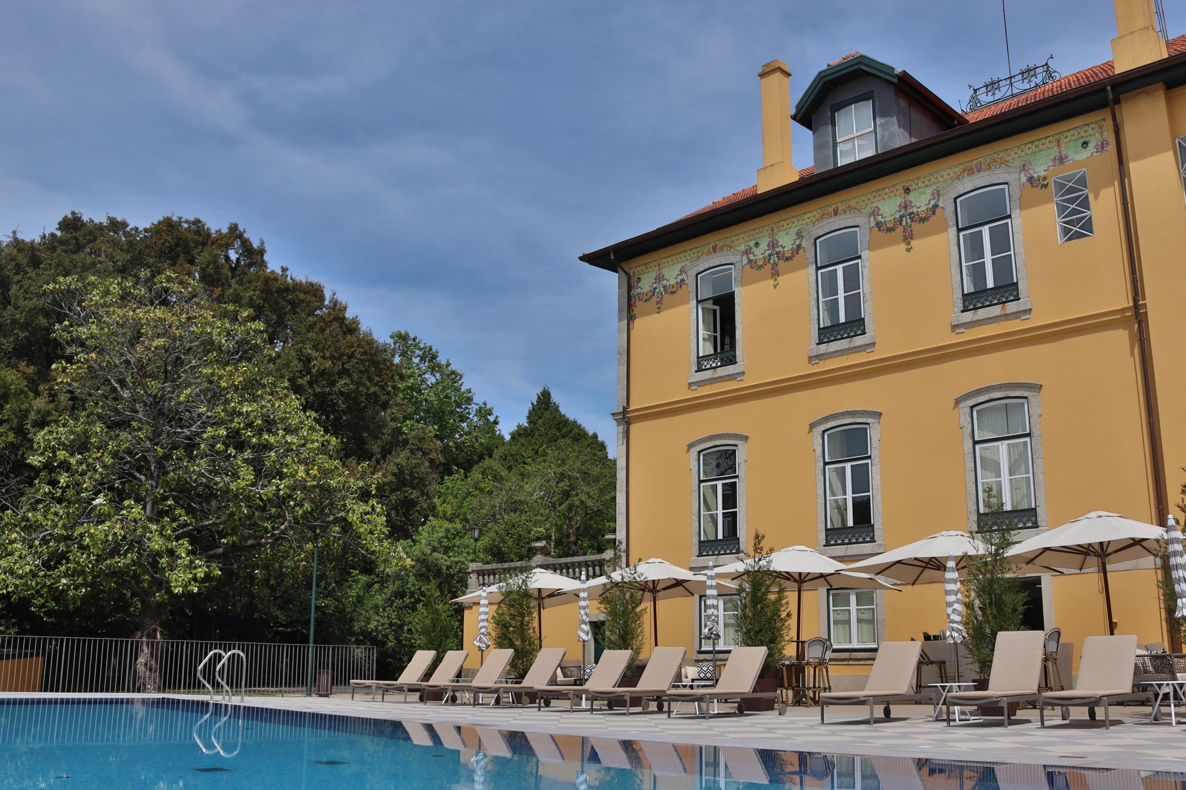 A poolside area with lounge chairs and umbrellas, a yellow building with decorative window frames, and a tree-lined background.