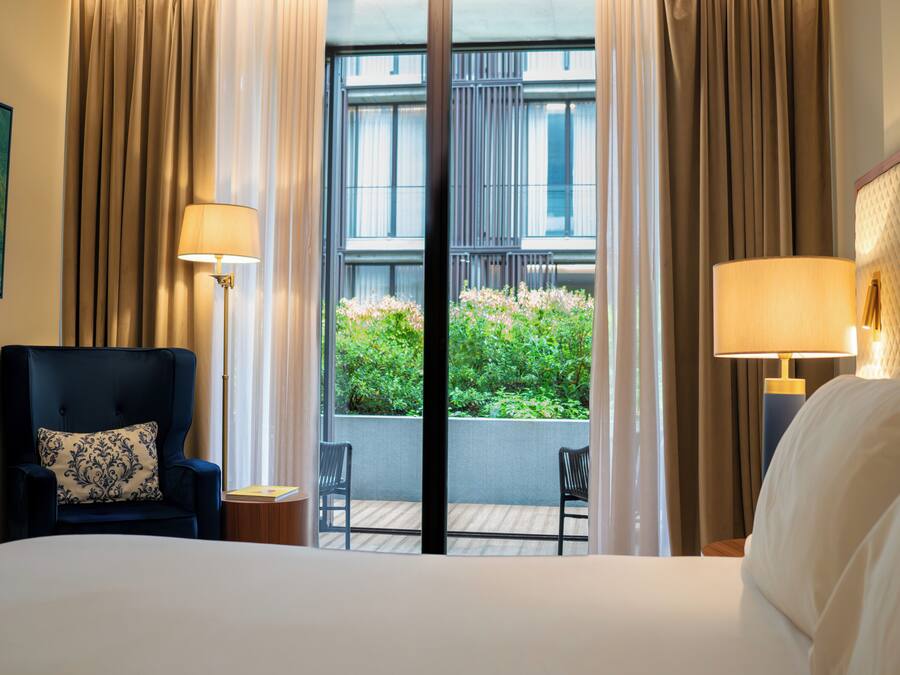 A hotel room with a bed, a chair, a table, and a view of greenery through large windows.
