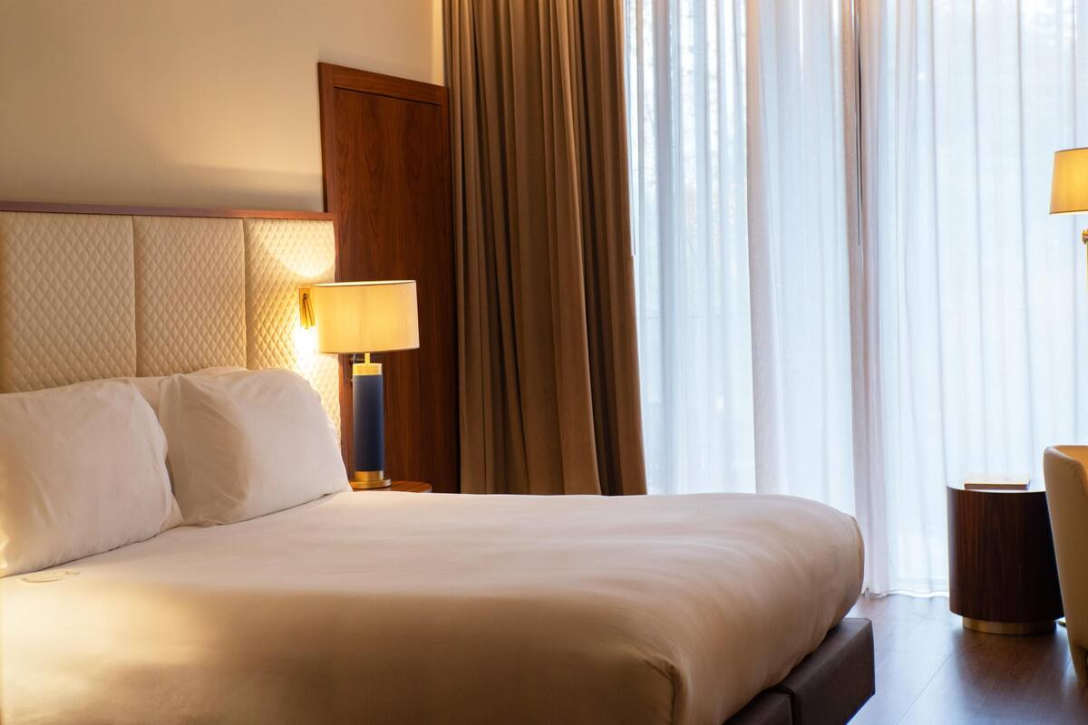A hotel room with a large bed, a nightstand with a lamp, a wooden headboard, and a view of the outdoors through sheer curtains.