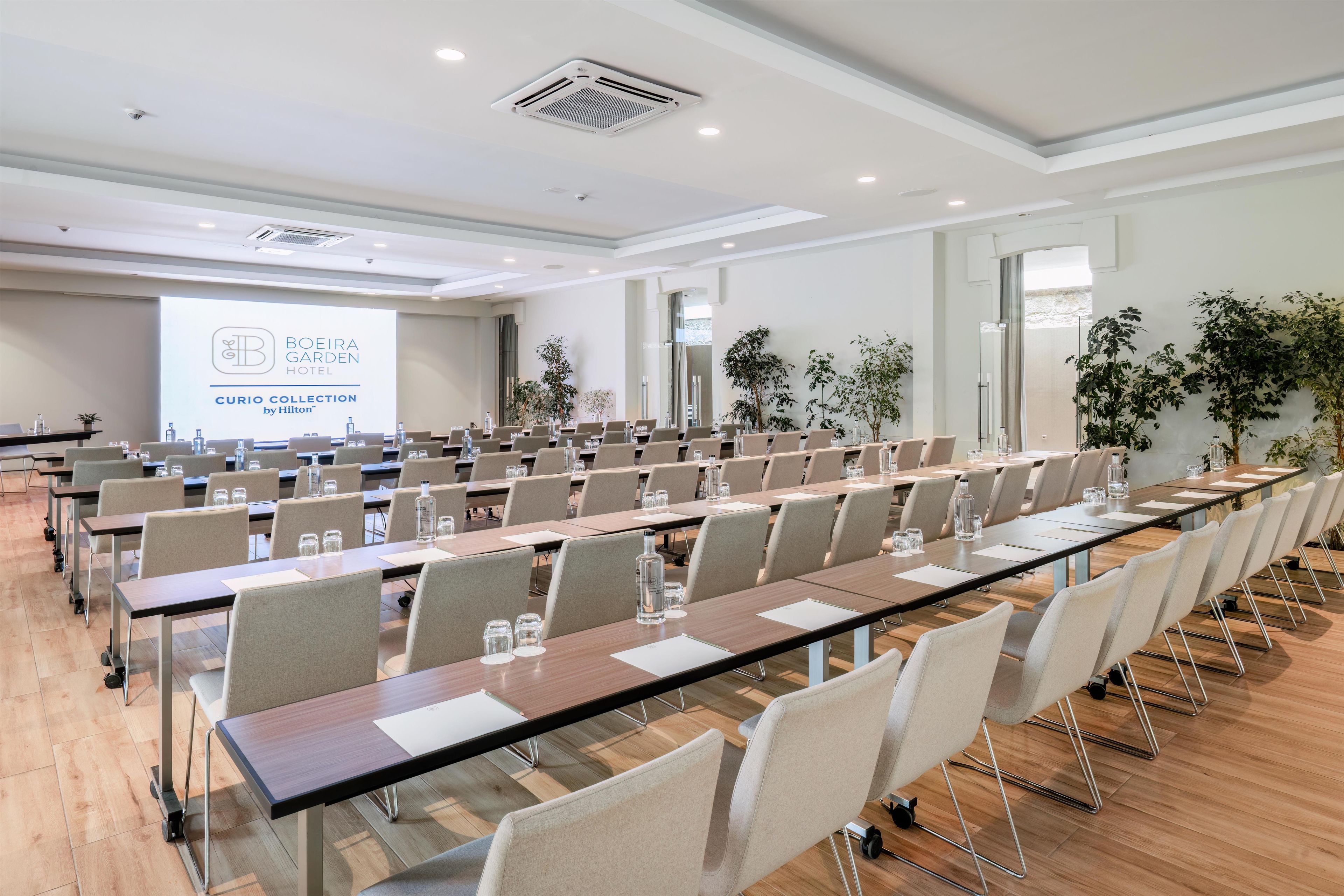 Ein Konferenzraum mit mehreren Tischen und Stühlen, einer Projektionsleinwand mit Hotelmarke und Zimmerpflanzen.