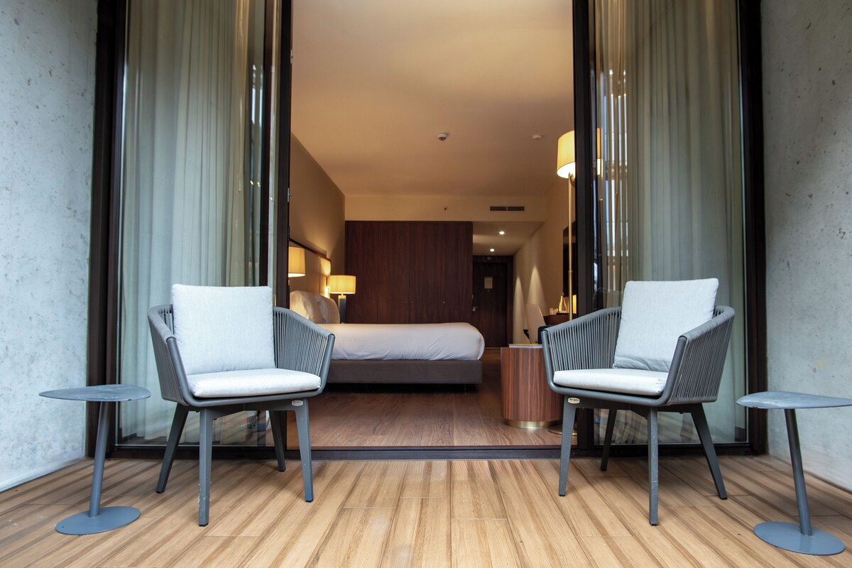 A hotel room with a bed, two armchairs, and two small tables.