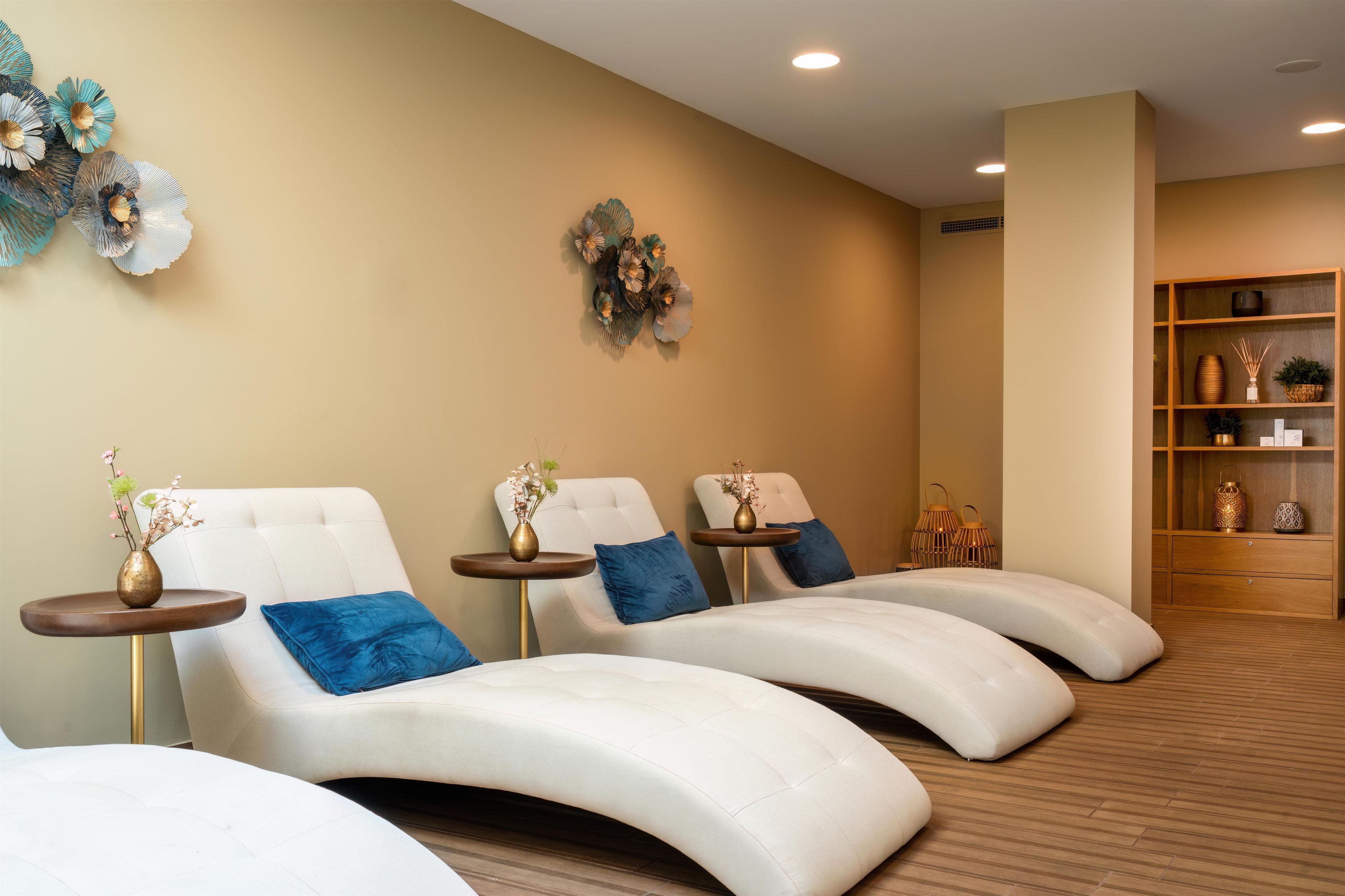 A spa room with three reclining chairs, a small table with a vase, and a shelf with decorative items.