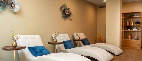 A spa room with three reclining chairs, a small table with a vase, and a shelf with decorative items.