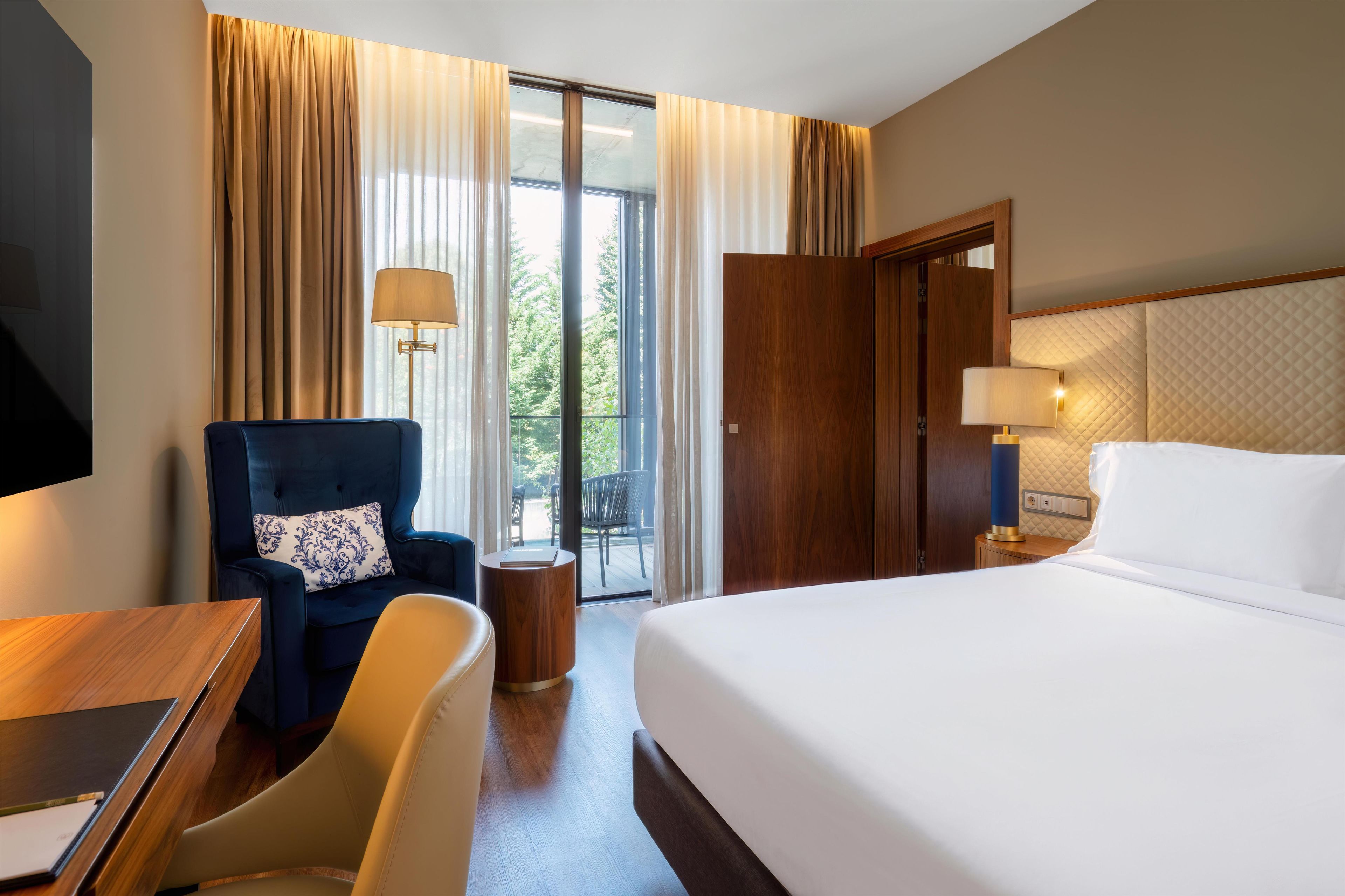 A hotel room with a bed, a desk, a chair, a lamp, and a view of the outdoors.