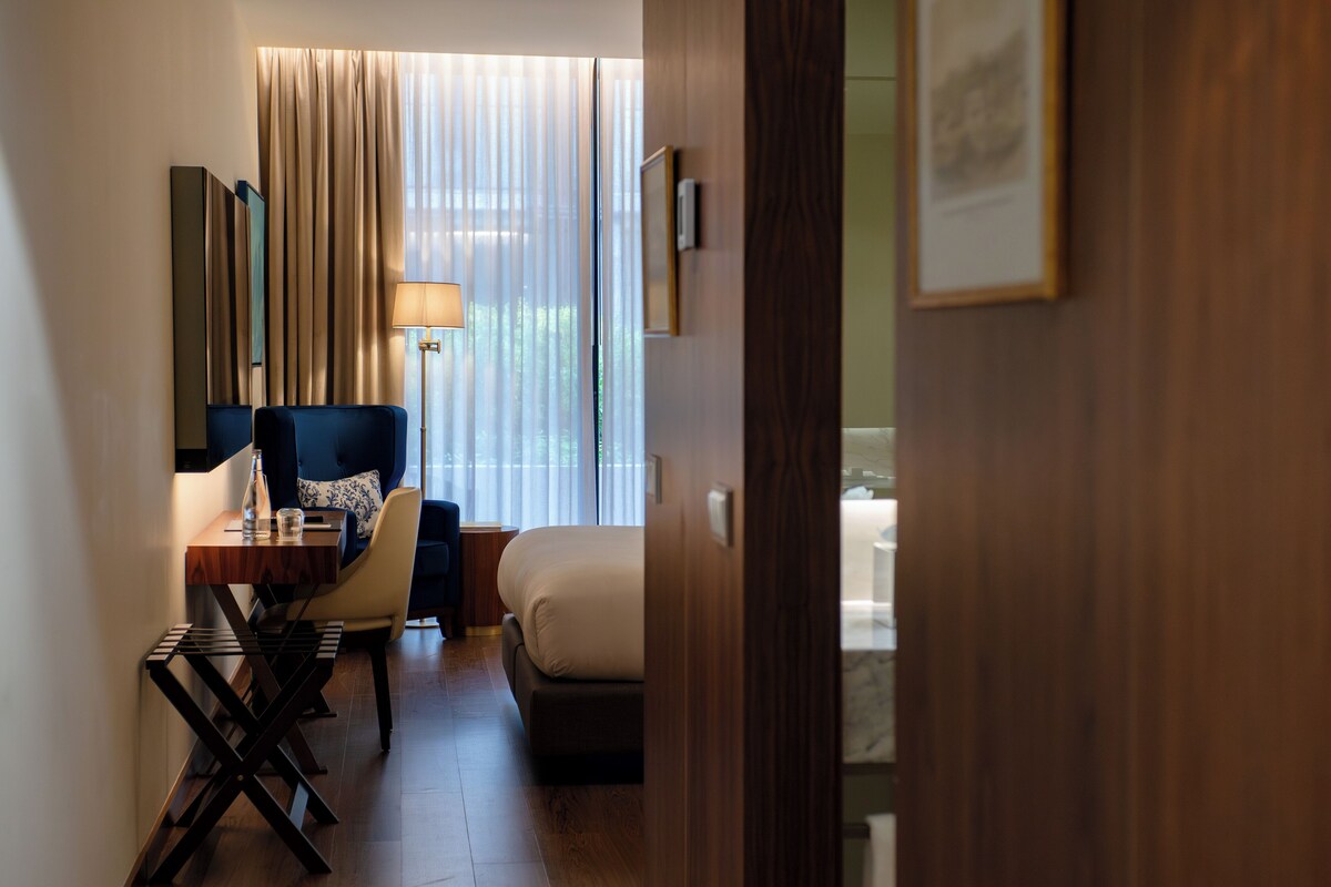 A hotel room with a bed, a desk with a chair, a lamp, and a window with curtains.