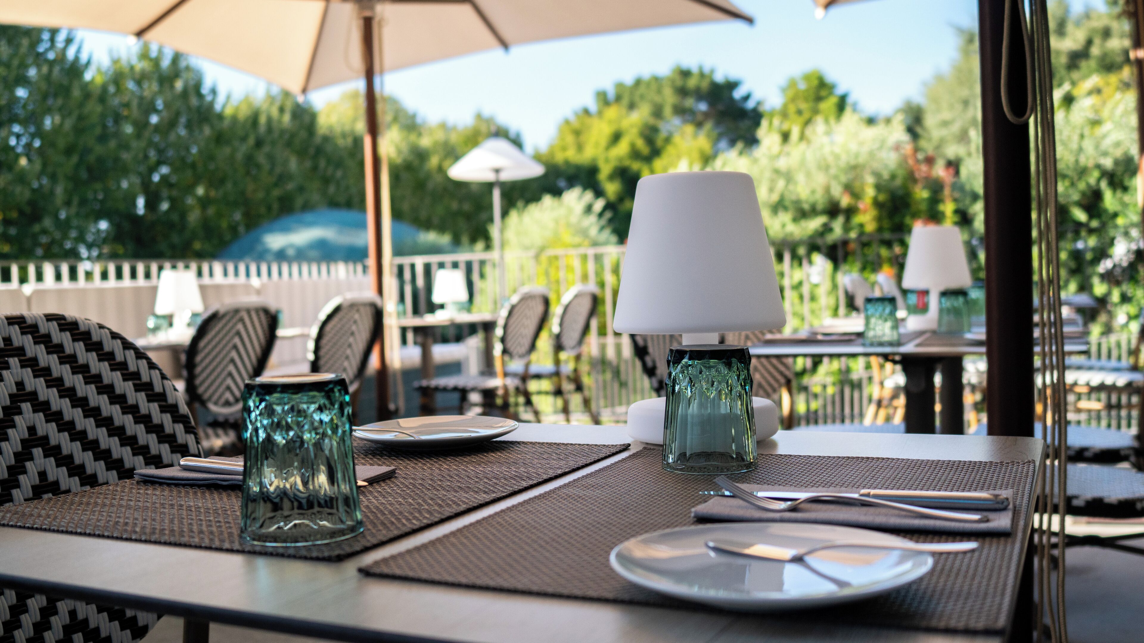 Un espace de restauration en plein air avec des tables préparées pour deux personnes, agrémenté de sets de table en fibres tressées, de vases en verre transparent et de lampes de table blanches.