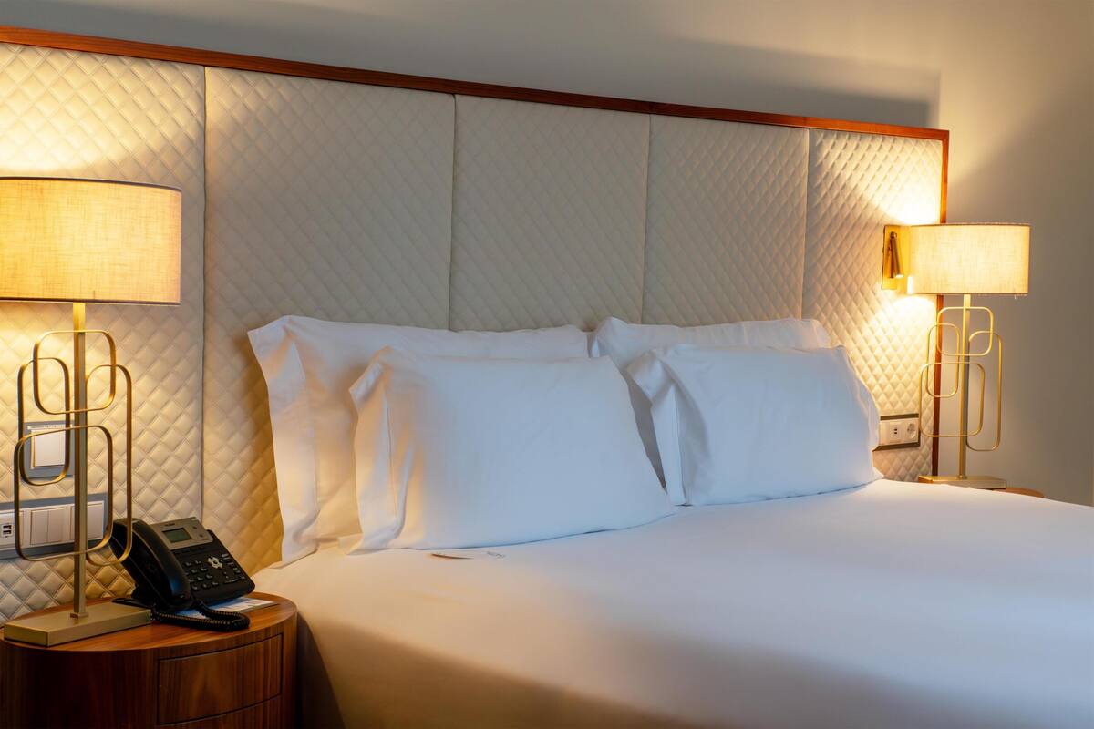 A hotel room with a bed, two bedside lamps, a telephone, and a wall with a quilted pattern.