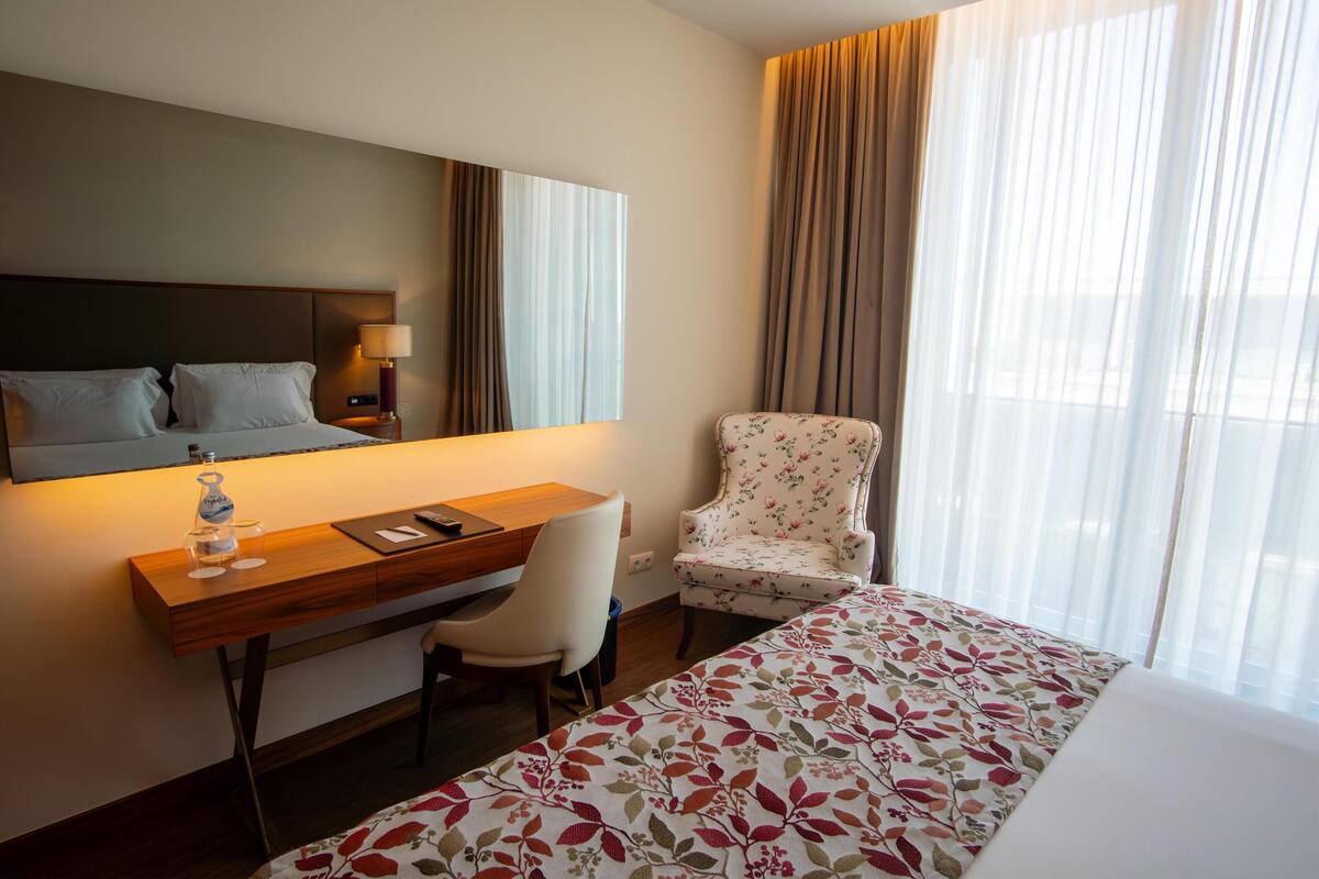 A hotel room with a bed, a desk, a chair, a lamp, and a large window with curtains.
