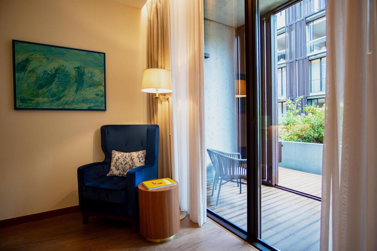A room with a dark blue armchair, a small wooden side table, a floor lamp, a painting on the wall, and a view of a balcony with a chair and greenery.
