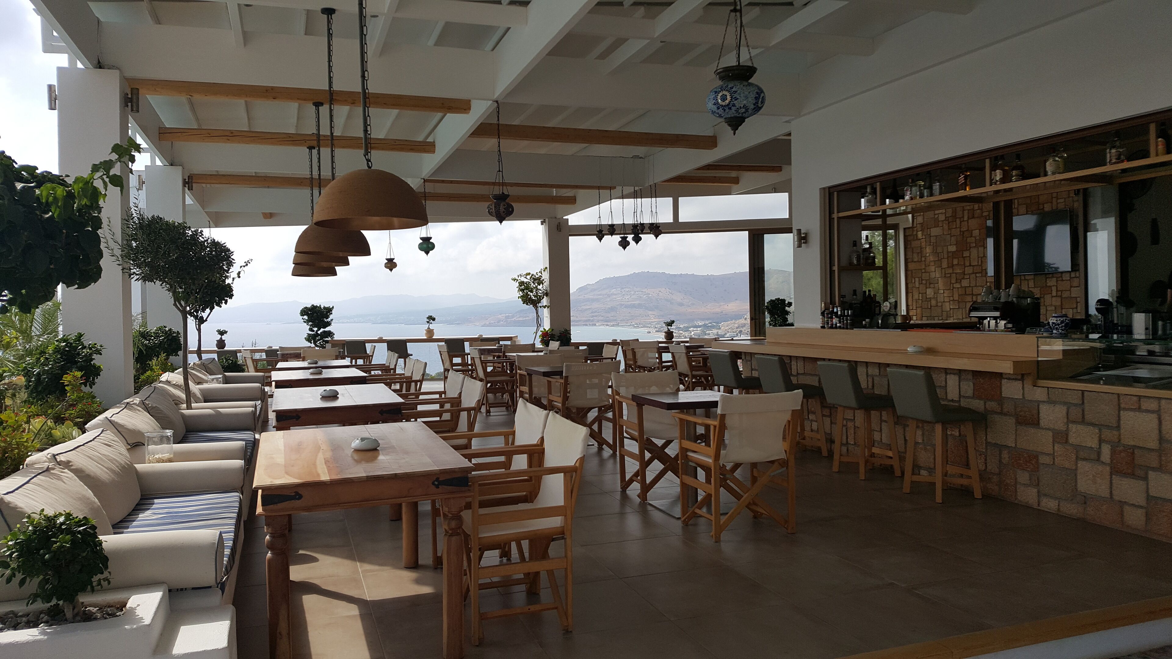 A spacious outdoor dining area with multiple tables and chairs, a bar area with a stone counter, and a view of the sea and mountains.