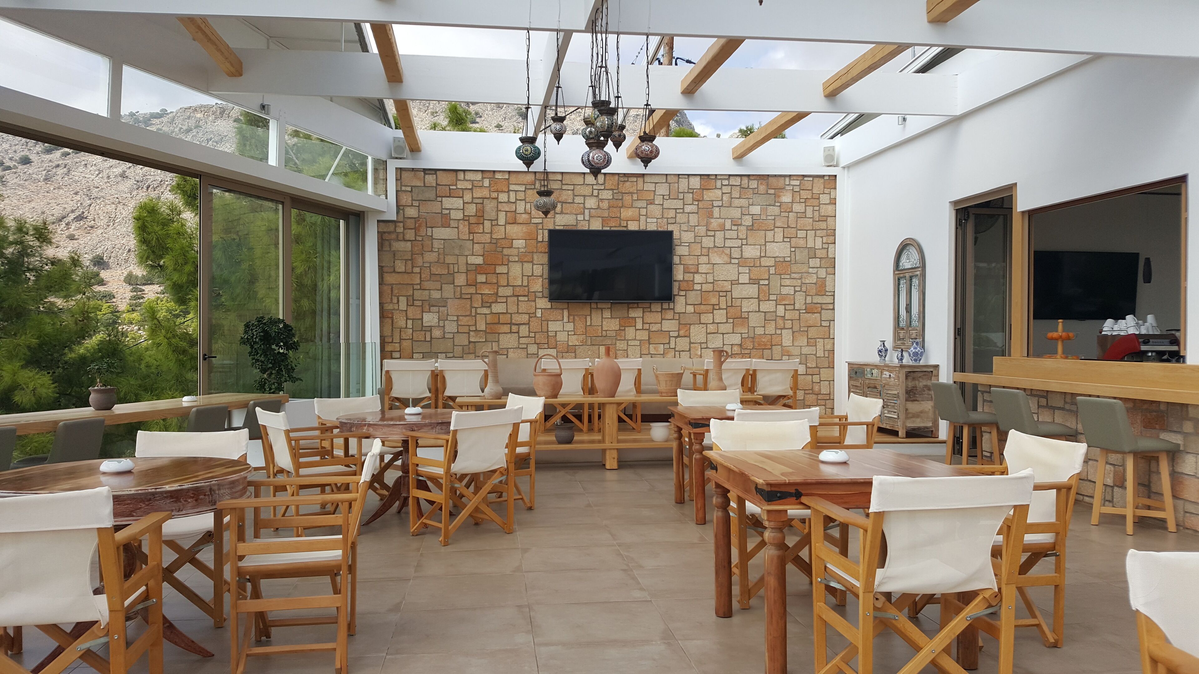 Un amplio comedor al aire libre con mesas y sillas de madera, una pared de piedra y un televisor montado arriba.