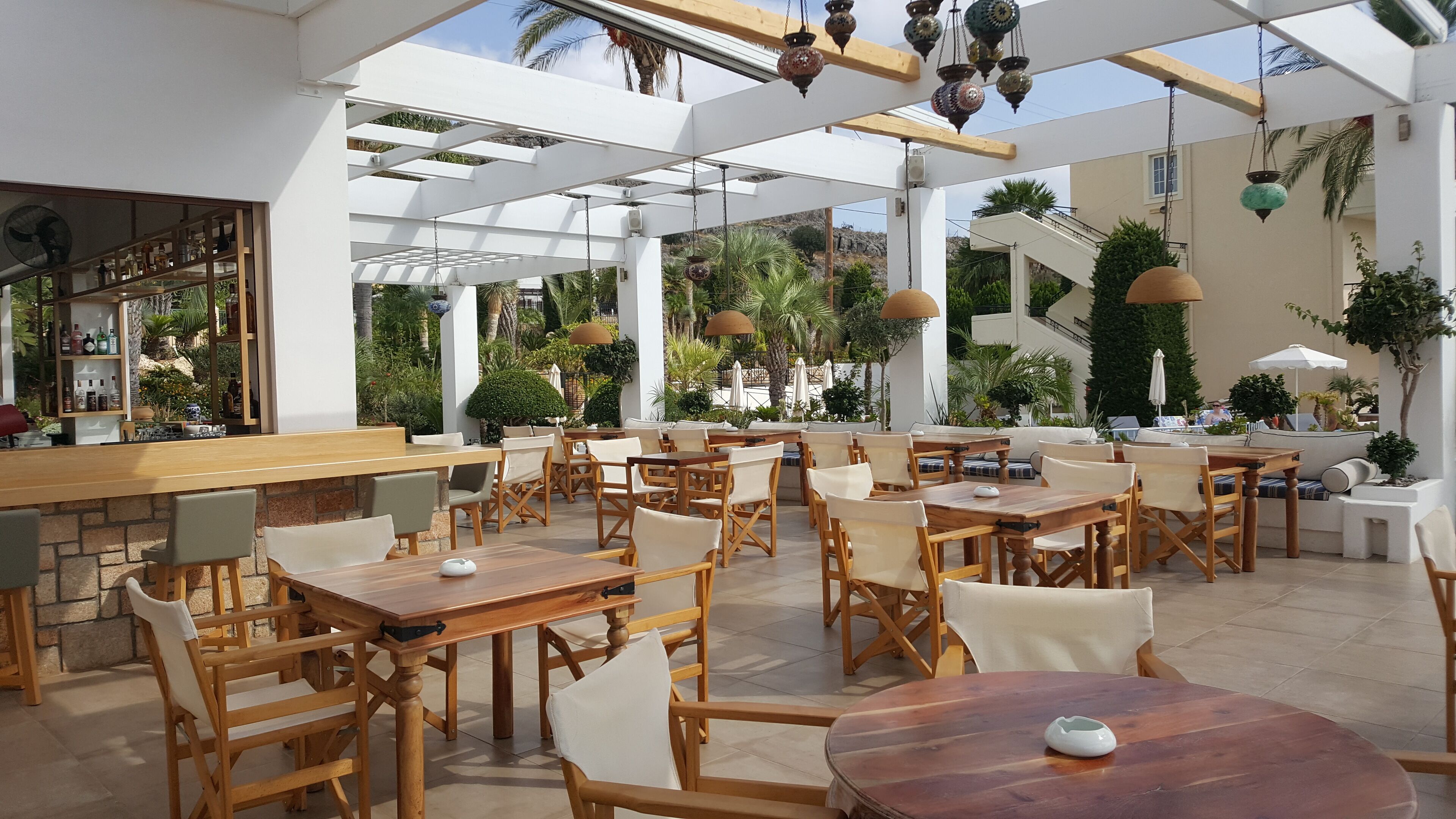 Un café al aire libre espacioso con mesas y sillas de madera, una zona de bar y luces decorativas colgantes.