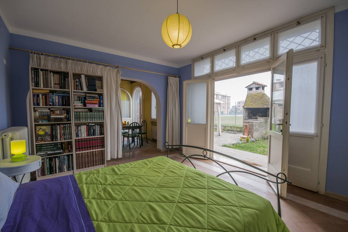 A bedroom with a green bed, a bookshelf, a small table with a lamp, and a view of the outside through a large window.