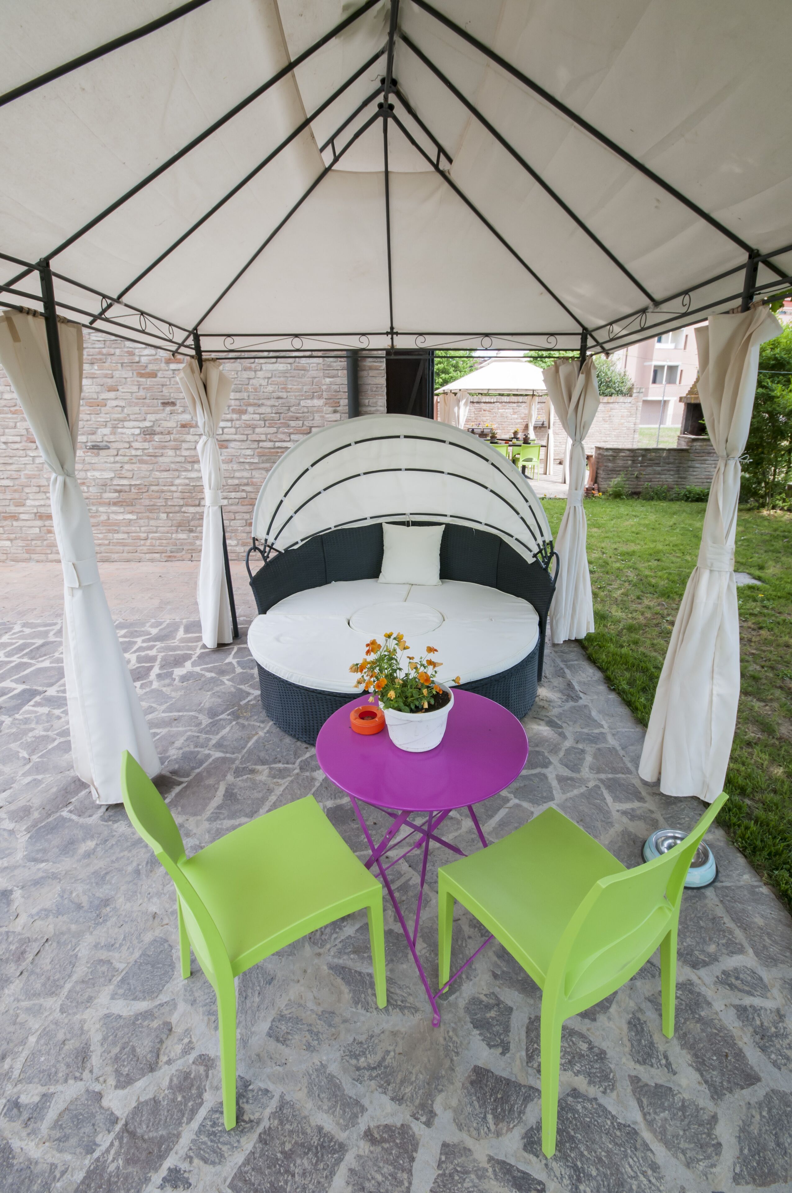 A covered outdoor seating area with a round sofa and two green chairs, a small table with a flower vase, and a stone floor.