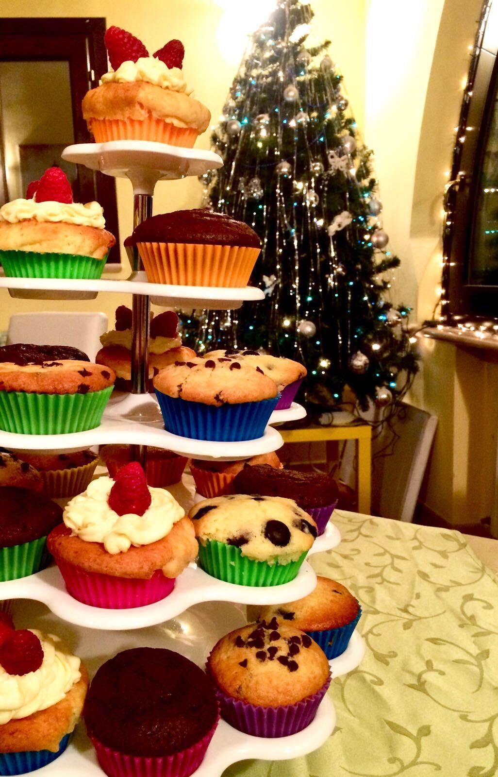 A three-tiered stand with various cupcakes and muffins, a Christmas tree in the background, and a patterned tablecloth.