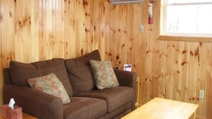 A wooden-paneled room with a brown sofa, two patterned cushions, a wooden table, and a fire extinguisher mounted on the wall.