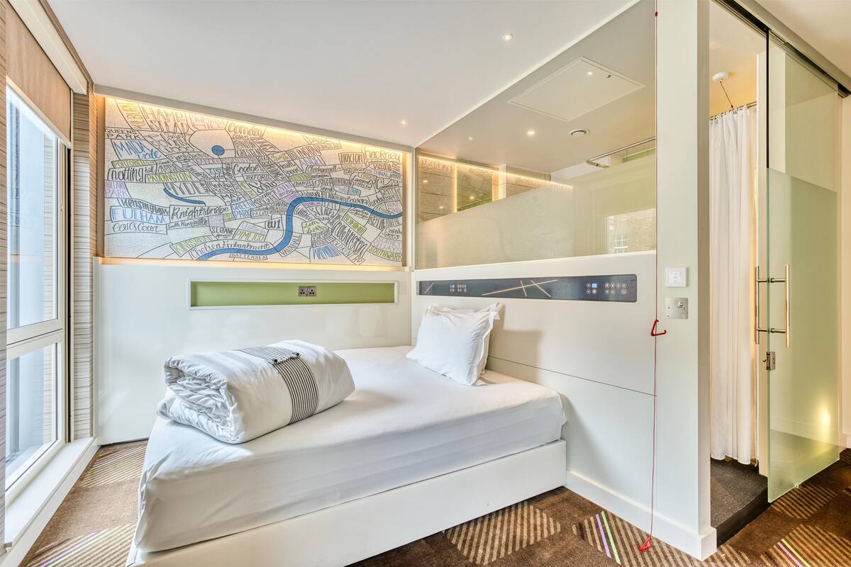 A modern hotel room with a large bed, a map on the wall, a glass partition, and a view of the city.
