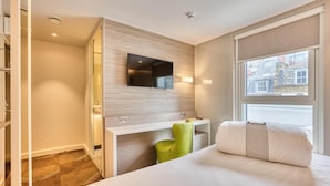 A hotel room with a bed, a desk, a green chair, a TV mounted on the wall, and a window with a view of buildings. - Hub By Premier Inn London - Soho (London)