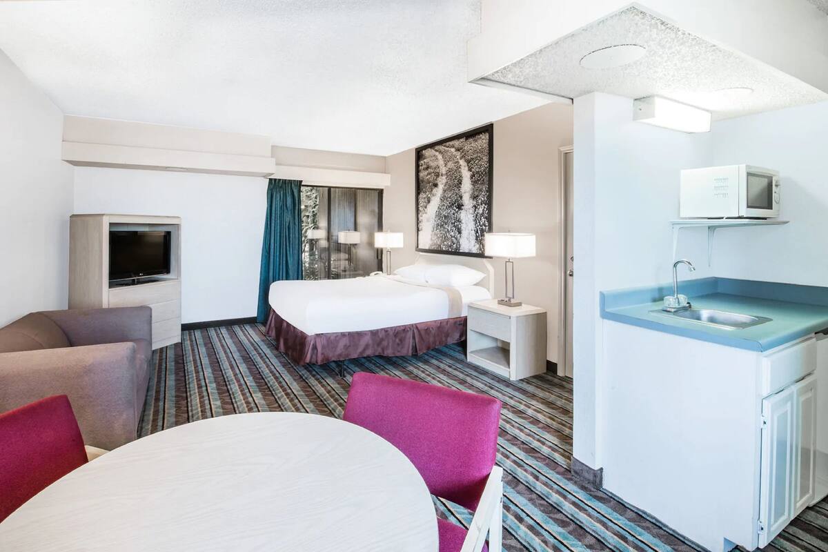 Habitación de hotel con cama, televisión, escritorio con computadora, silla, mesa y baño con lavamanos y espejo.