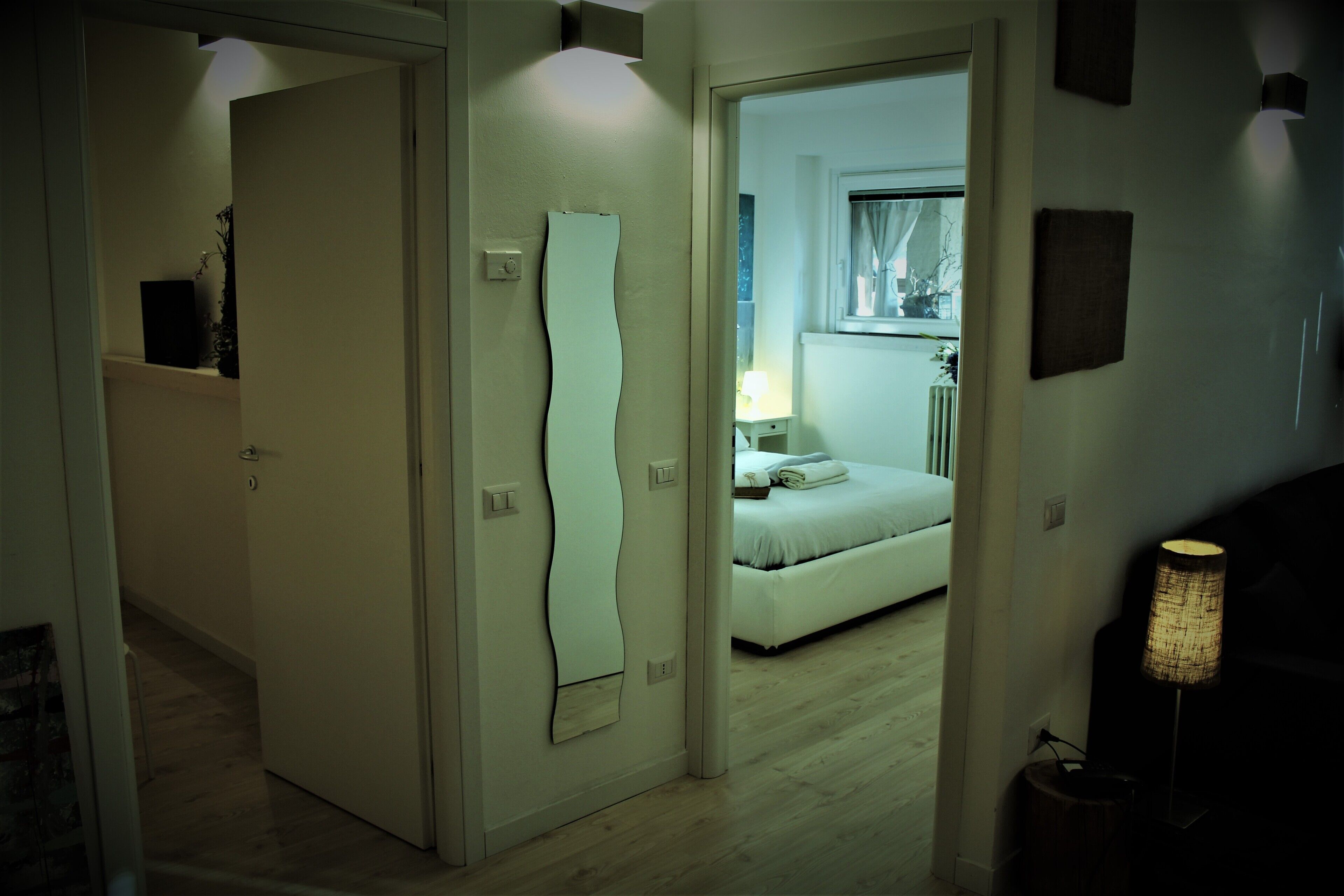 A hallway with a door leading to a bedroom. The bedroom has a bed, a nightstand, and a lamp.